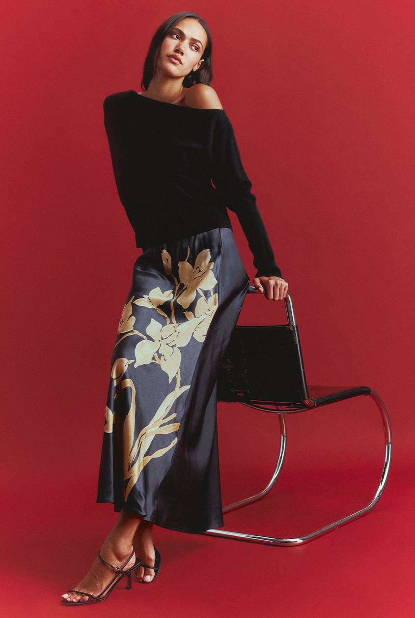 Woman in a black off-the-shoulder sweater and a navy satin midi skirt with a large gold floral print, standing beside a chrome and black leather cantilever chair against a deep red backdrop.
