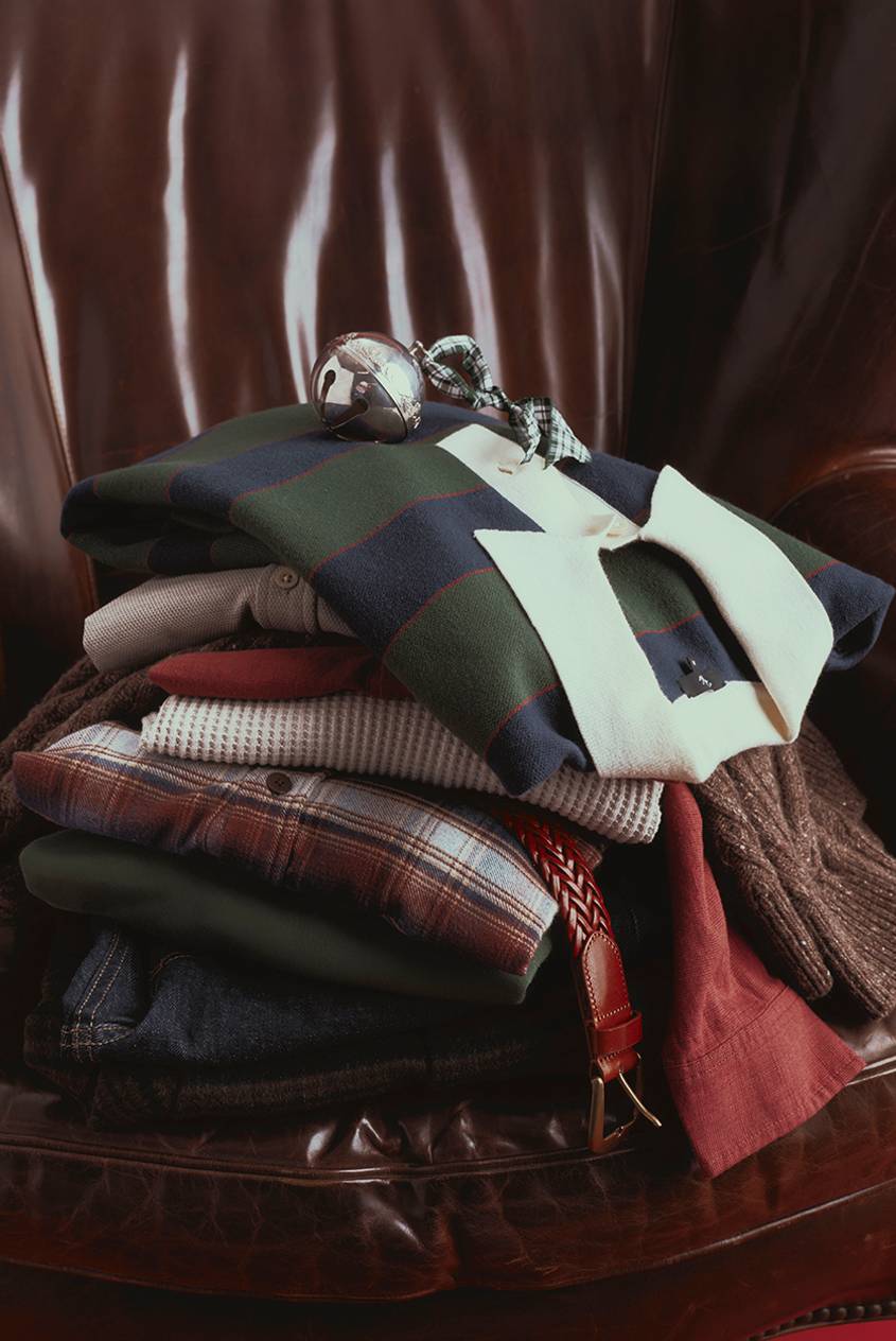 Stack of folded menswear on a brown leather armchair, topped by a navy and green striped rugby shirt with a cream collar and a silver sleigh bell tied with a green plaid ribbon. Below it are a waffle-knit top, a brown and red plaid shirt, a green sweater, dark denim, and a braided brown leather belt, with a rust red shirt and a brown tweed knit hanging off the sides.