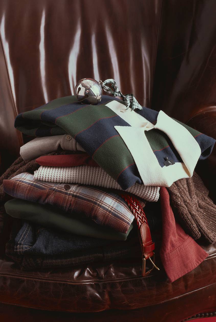 Stack of folded menswear on a brown leather armchair, topped by a navy and green striped rugby shirt with a cream collar and a silver sleigh bell tied with a green plaid ribbon. Below it are a waffle-knit top, a brown and red plaid shirt, a green sweater, dark denim, and a braided brown leather belt, with a rust red shirt and a brown tweed knit hanging off the sides.