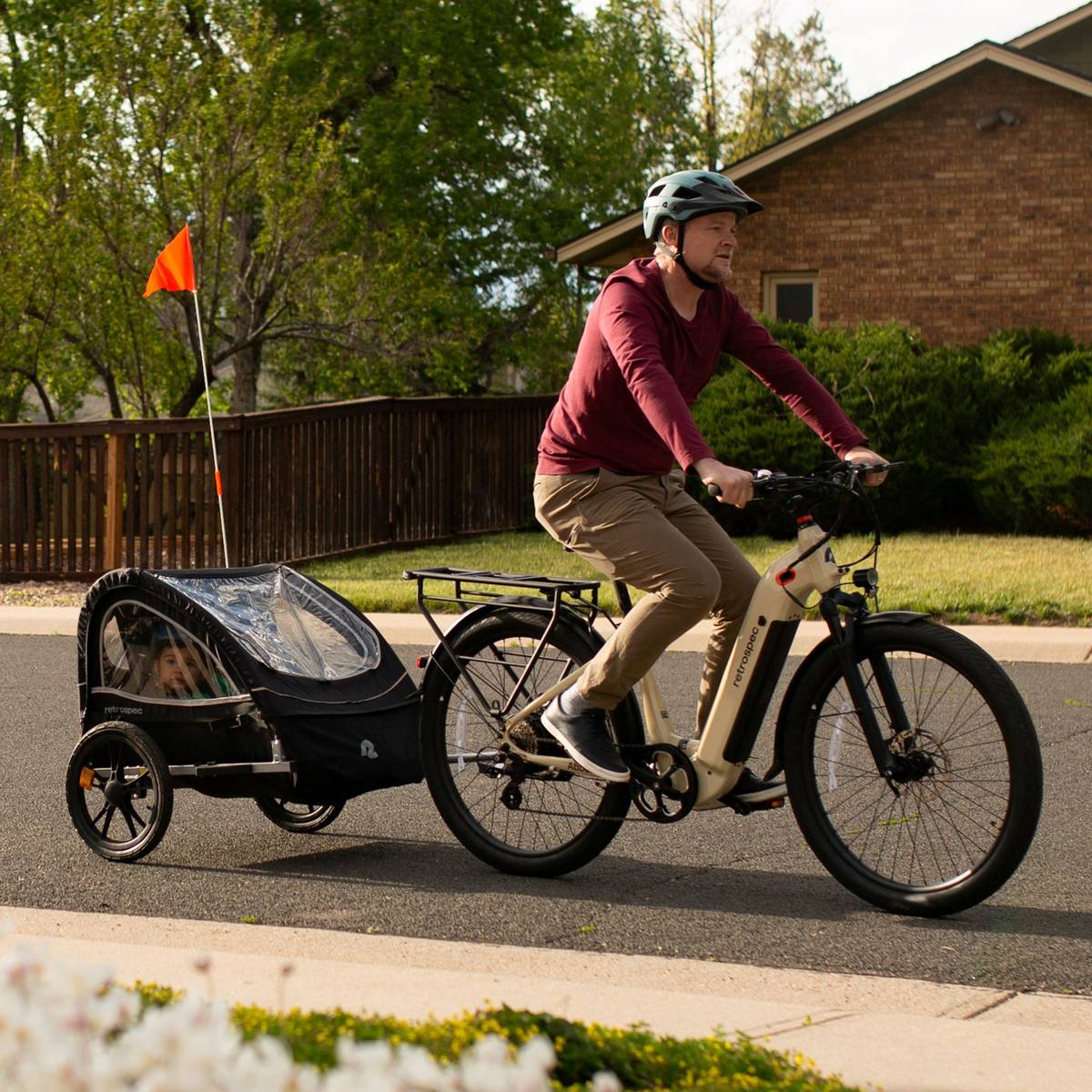 A cyclist in a maroon shirt rides an electric bike with a Rover Kids' Bike Trailer, set against a suburban backdrop of green trees and a house.