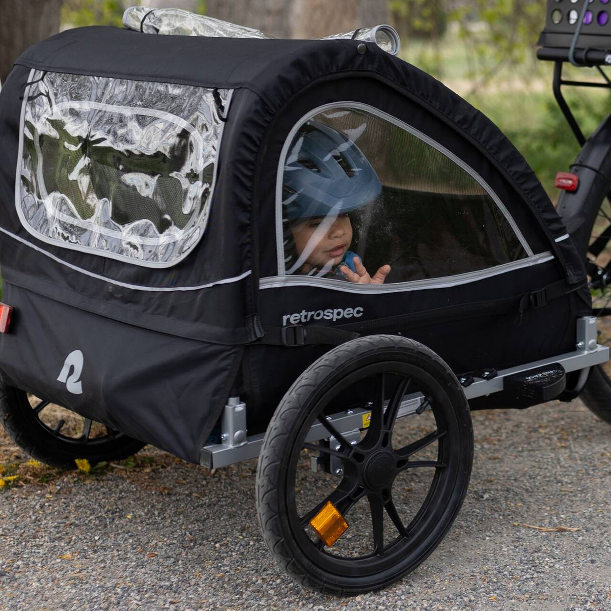 A black Rover Kids' Bike Trailer with a transparent front and wheels sits on a gravel path, showcasing its modern design and safety features.