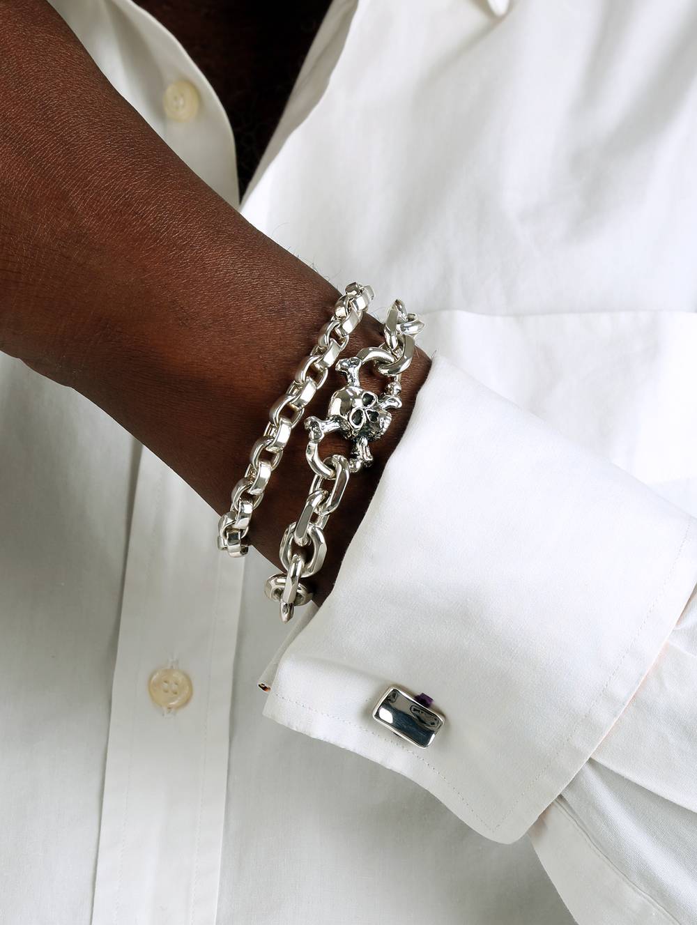 Man is wearing a white shirt with two chunky silver bracelets, one with a skull and cufflinks on his sleeve.