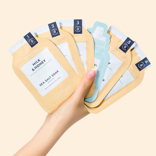 Hand Holding Pedi in a Box Ultimate 6 Step Milk & Honey Box Packets on Yellow Background