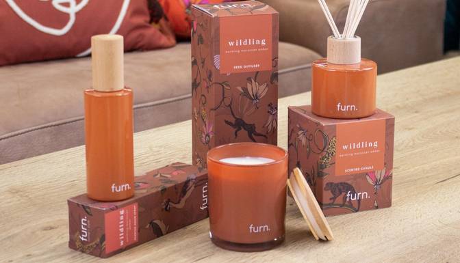 A selection of home fragrance products and their packaging, including a room spray, scented candle and reed diffuser, laid out on a wooden table in front of a neutral sofa.