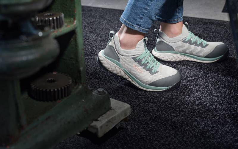 Knee down shot of woman wearing grey and granite-green Arvada Shift work sneakers and walking past metal tool shelf. 