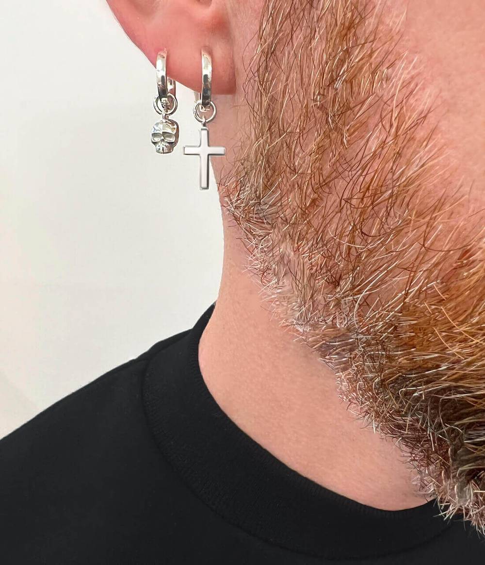 Man with black top and beard is wearing a silver cross hoop earring and a skull hoop earring in silver on the same ear
