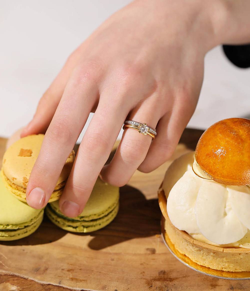 A woman's hand, who wears the 'Lotus' yellow gold diamond engagement ring and the 'Halo' platinum diamond wedding band is grabbing a macaron from a table full of desserts.