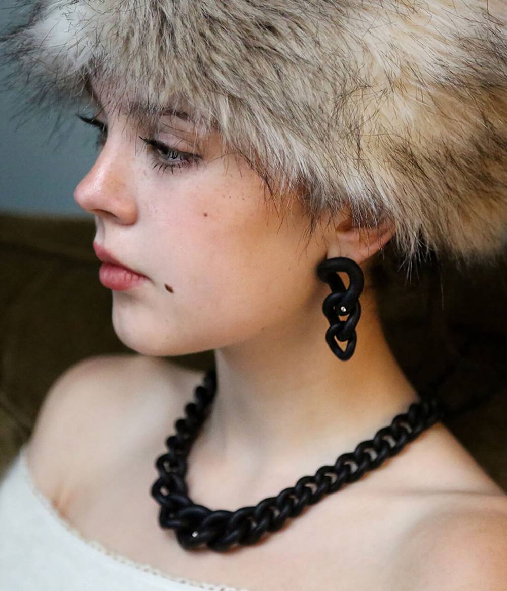 A girl is wearing a statement black chain necklace with a diamond and an black chain earring with a diamond from the Raven collection.