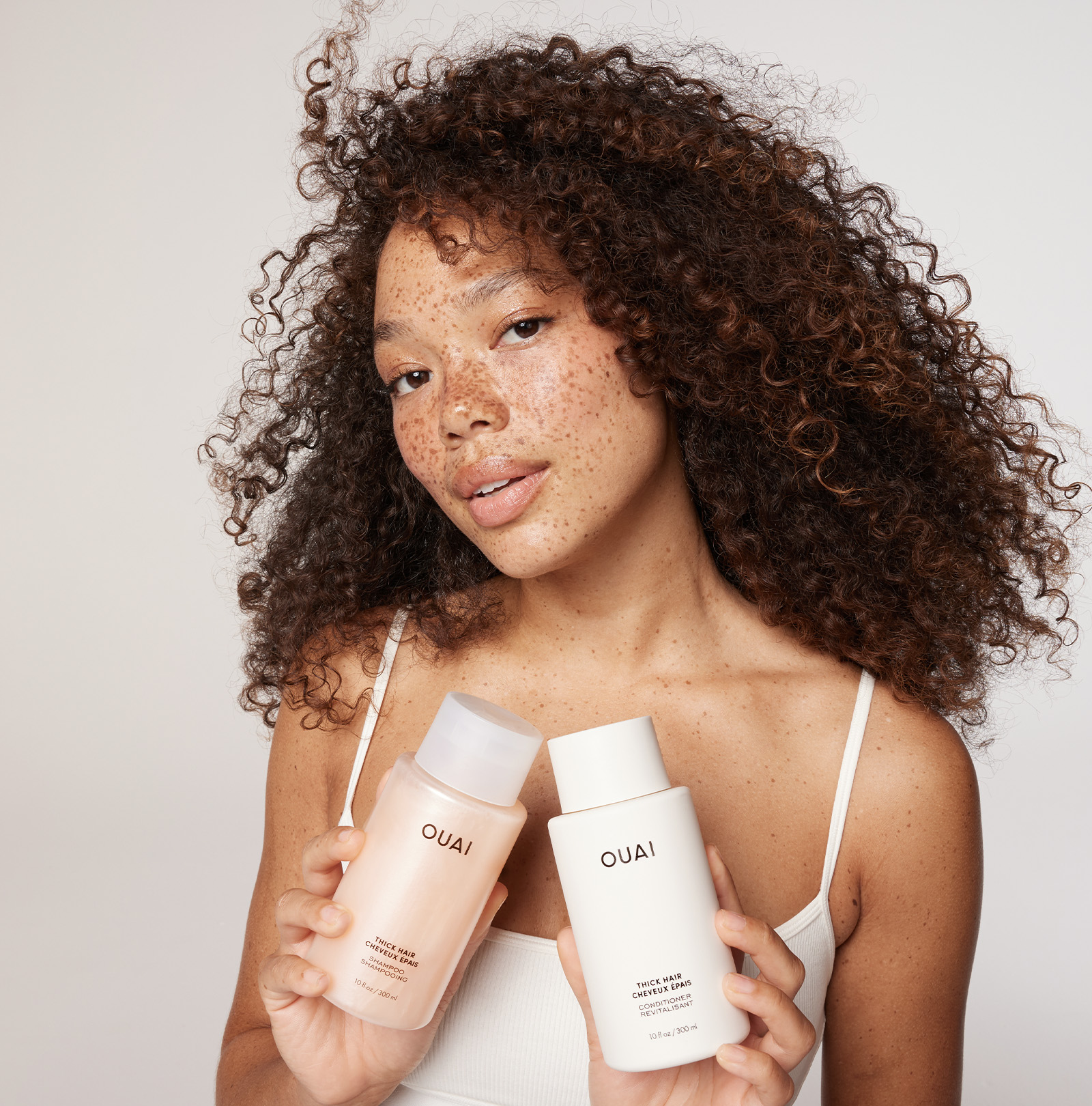 A woman with voluminous curly hair holds two OUAI hair care products in front of her. She is wearing a white tank top. One bottle is a peach-colored shampoo, and the other is a white conditioner.