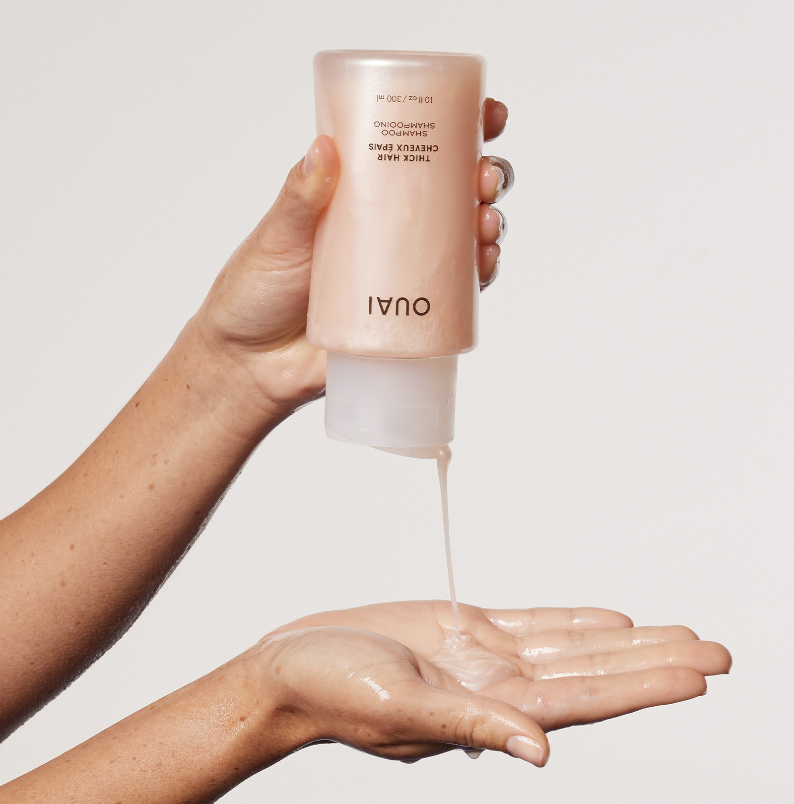 A close-up of a person dispensing OUAI Thick Hair Shampoo into their palm. The peach-colored bottle is held upside down, and a creamy stream of shampoo is flowing into an open hand.