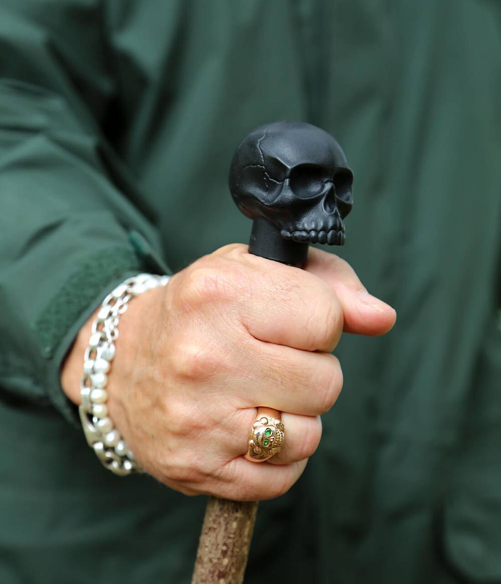 A man who is wearing a silver and a pearl bracelet, as well as a rose gold smiling skull ring is holding a hazel wood walking stick with a  skull caster seed oil black top.