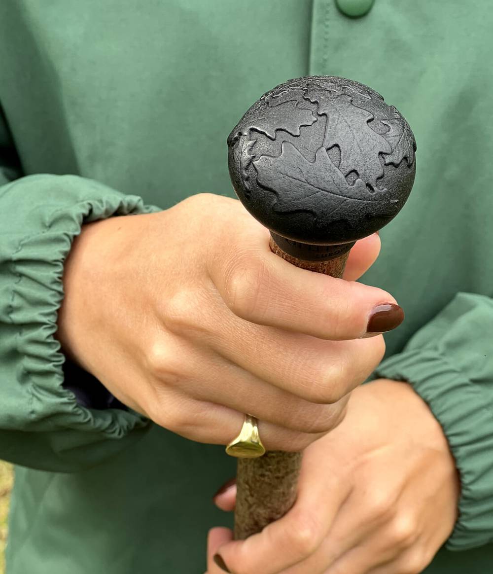 Woman is holding a caster seed oil and hazel wood walking stick and wearing an oval yellow gold signet ring.