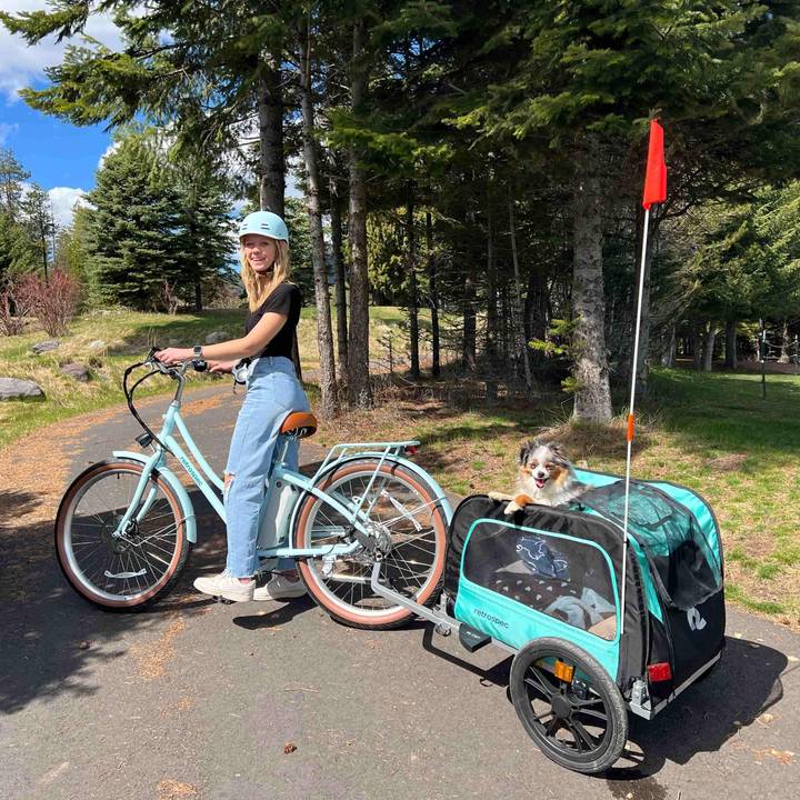 A smiling woman in a light blue helmet and jeans stands with her retrospec cruiser bike on a sunny paved path surrounded by tall pine trees. A happy tri-color Australian Shepherd peeks out of a teal and black retrospec pet trailer hitched to the back of the bike, ready for the ride.