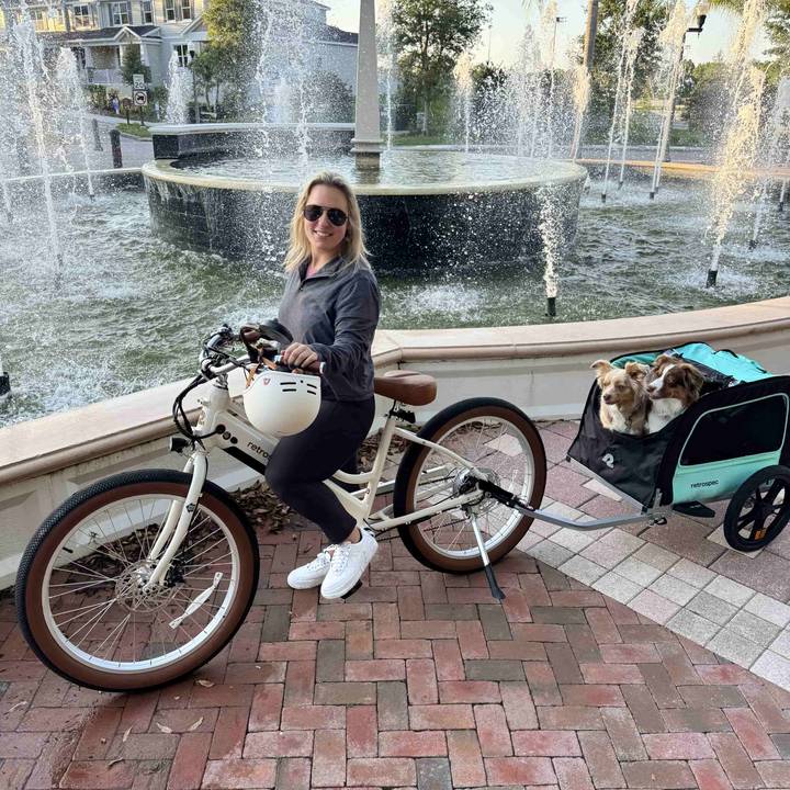 A woman in sunglasses and a gray pullover sits on a cream-colored retrospec EBike with brown accents, smiling in front of a large decorative fountain. Two Australian Shepherds ride comfortably in a teal and black retrospec pet trailer hitched behind the bike, all set for a sunset cruise.