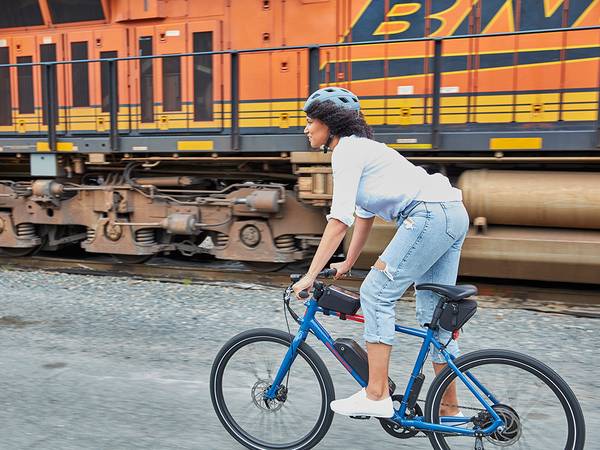 Woman riding a blue electric bike with a train in the background.