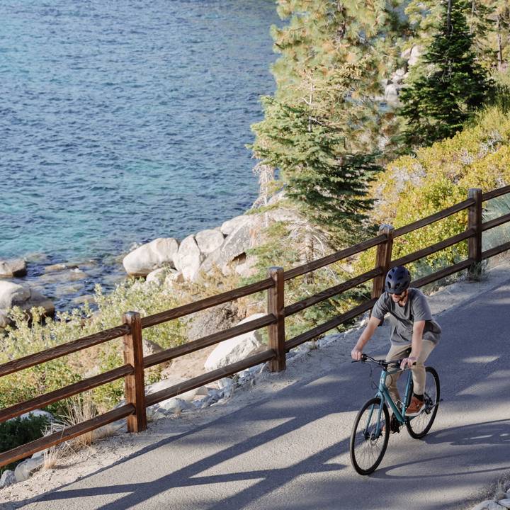 A person rides a bicycle along a scenic pathway near a lake, surrounded by lush trees and rocky shores under bright sunlight.