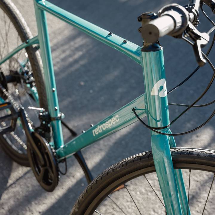 A close-up of a teal Atlas Fitness 21 Speed Hybrid Bicycle's frame, showcasing its sleek design and logo against a sunlit, paved surface.