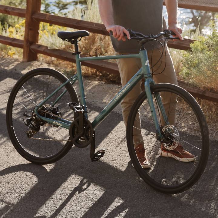A person stands beside a teal Atlas Fitness Hybrid 21 Speed Bike on a path, with a wooden fence and greenery in the background.