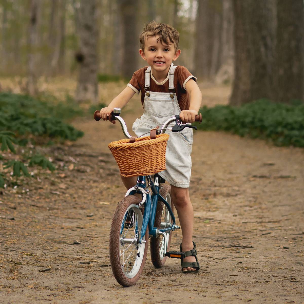 A young child in overalls rides a blue Beaumont Mini 16" Kids' Bike with a woven basket down a tree-lined path in a forested area.