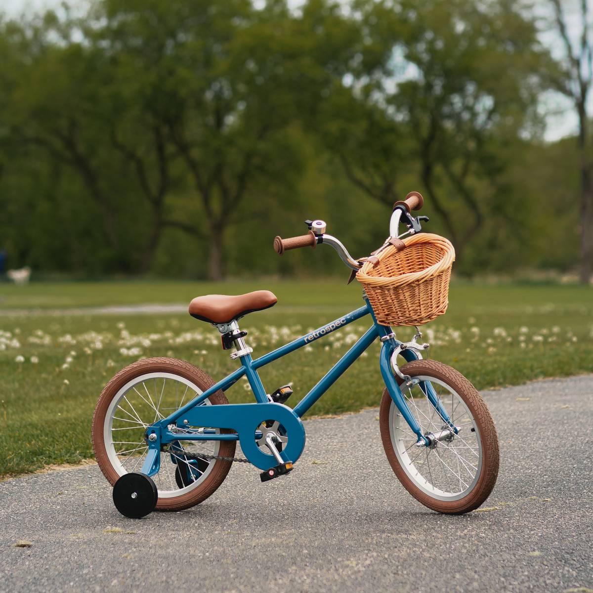 A blue Beaumont Mini 16" Kids' Bike with a wicker basket, brown handlebars, and training wheels parked on a pathway, surrounded by greenery.