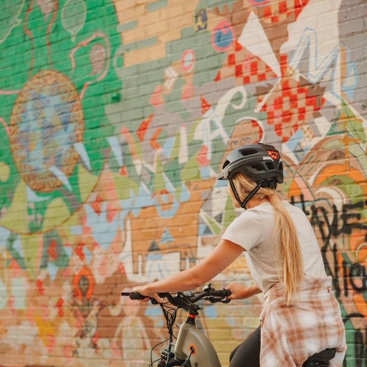 A woman rides a bike wearing a Lennon Commuter Bike Helmet in front of a colorful graffiti wall, showcasing urban art and vibrant street culture.