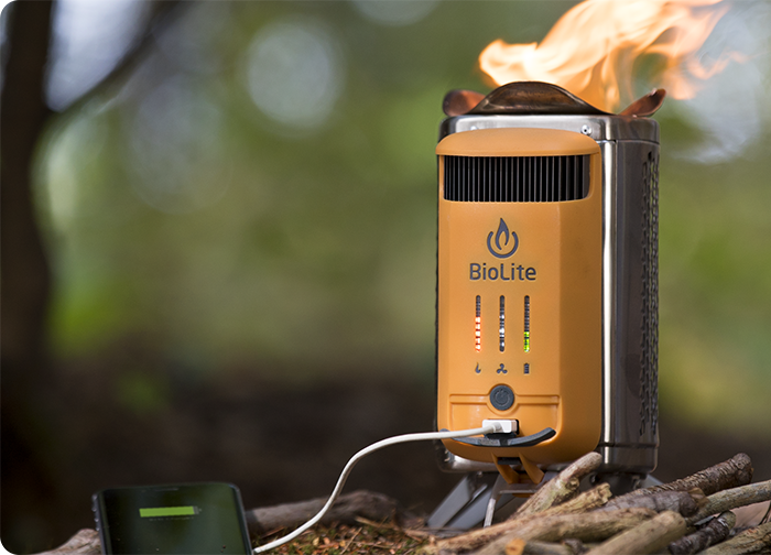 A BioLite camp stove with flames on top, charging a device, surrounded by sticks in a natural setting.