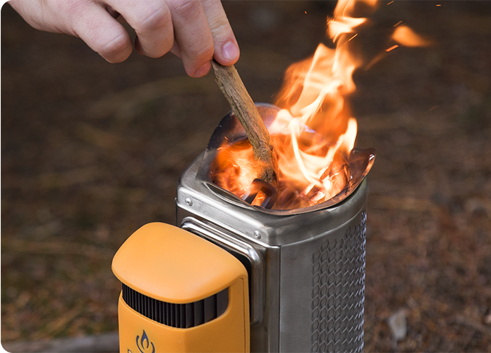 A hand adds a stick to a metal stove, where flames are rising, set against a natural background.