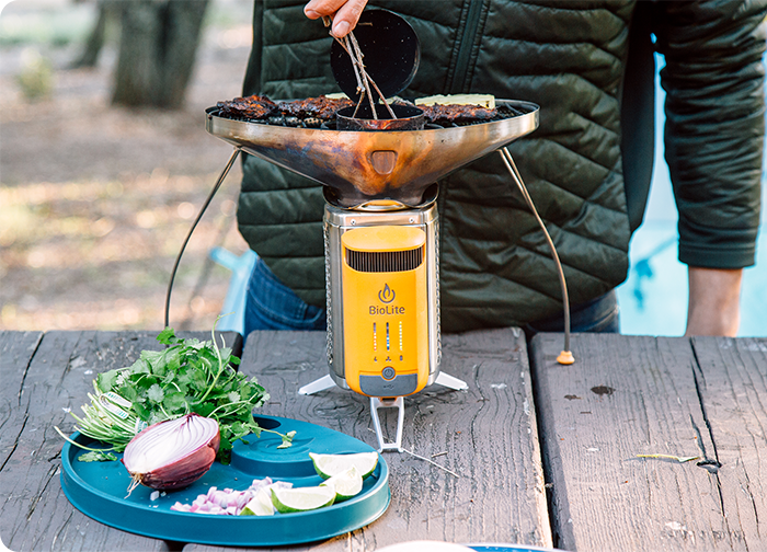 A person grilling food on a portable BioLite stove, with fresh ingredients like cilantro, onion, and lime on a blue plate nearby.