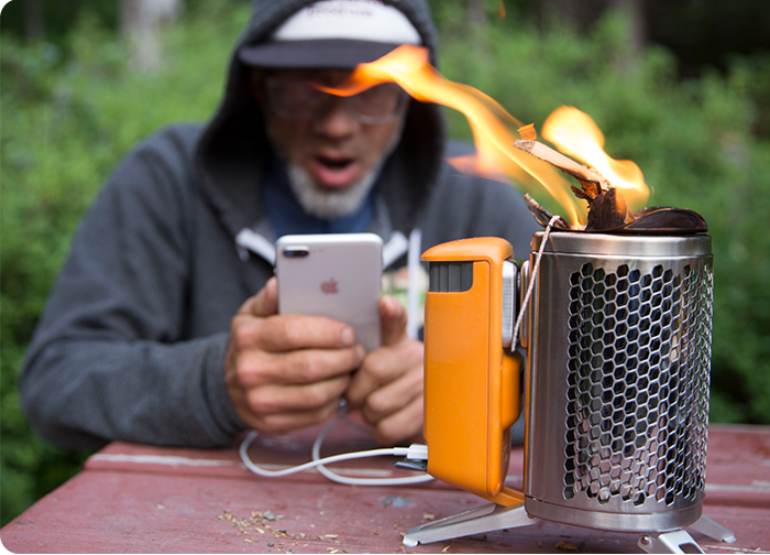 A person in a hoodie is surprised while using a phone, next to a portable stove with flames and an orange accessory.