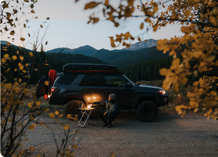 A person sits at a table beside a parked SUV in a forested area, surrounded by autumn leaves and mountains at dusk.