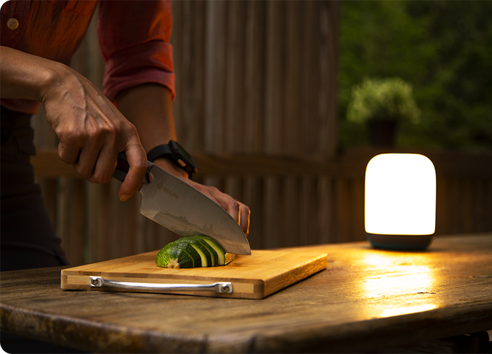A person slices a cucumber on a wooden cutting board, illuminated by a soft light, with a rustic background.