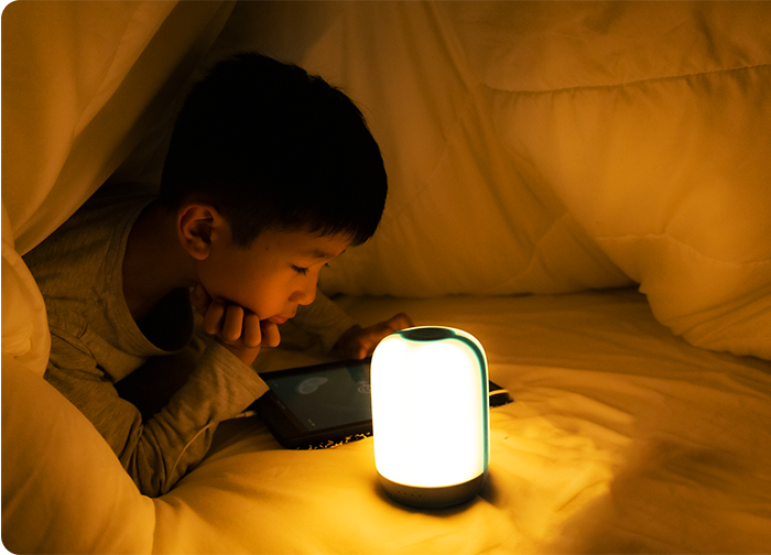A child is lying under a blanket, focused on a tablet, with a soft glowing lamp beside them.