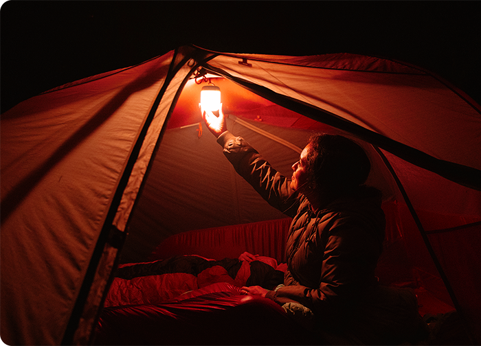 A person inside a tent holds a lantern, casting a warm glow in the dark. The tent's interior is cozy and inviting.