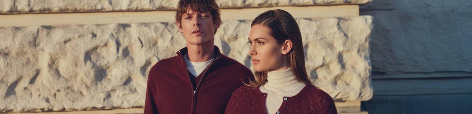 Man and woman standing outdoors against a stone wall, both wearing burgundy knitwear — the man in a zip-neck sweater over a white T-shirt, and the woman in a buttoned cardigan over a cream turtleneck