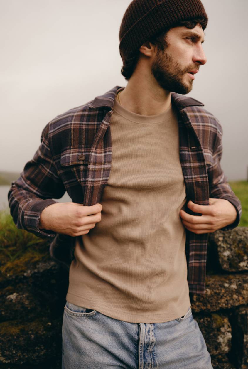 Model wearing a brown and purple plaid overshirt over a beige ribbed T-shirt with light wash jeans, shown outdoors.