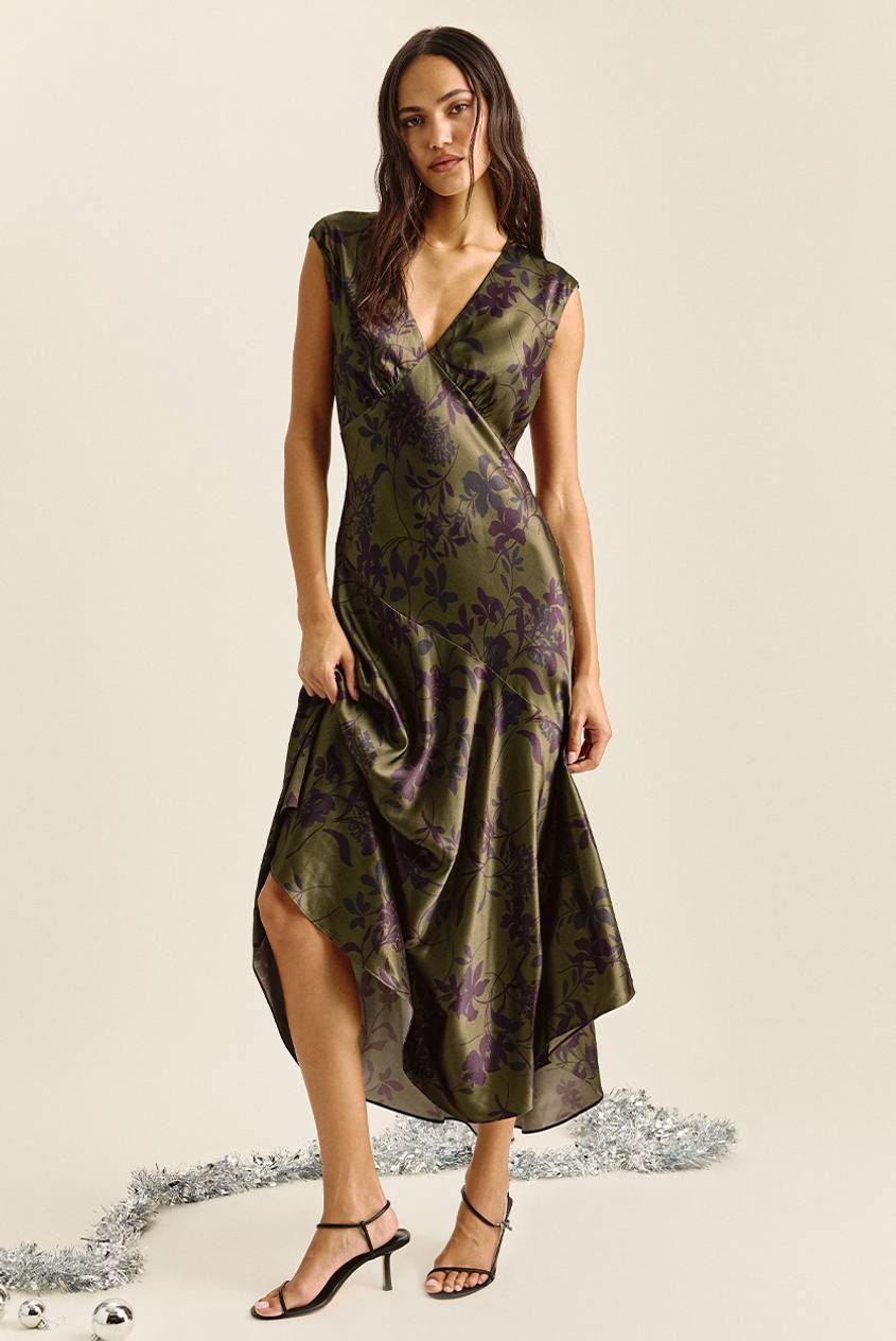 Woman standing and holding the skirt of a sleeveless olive green satin midi dress with a deep V-neck, a purple floral print, and an asymmetrical hem, paired with black strappy heels. Silver tinsel and ornaments lie on the floor.
