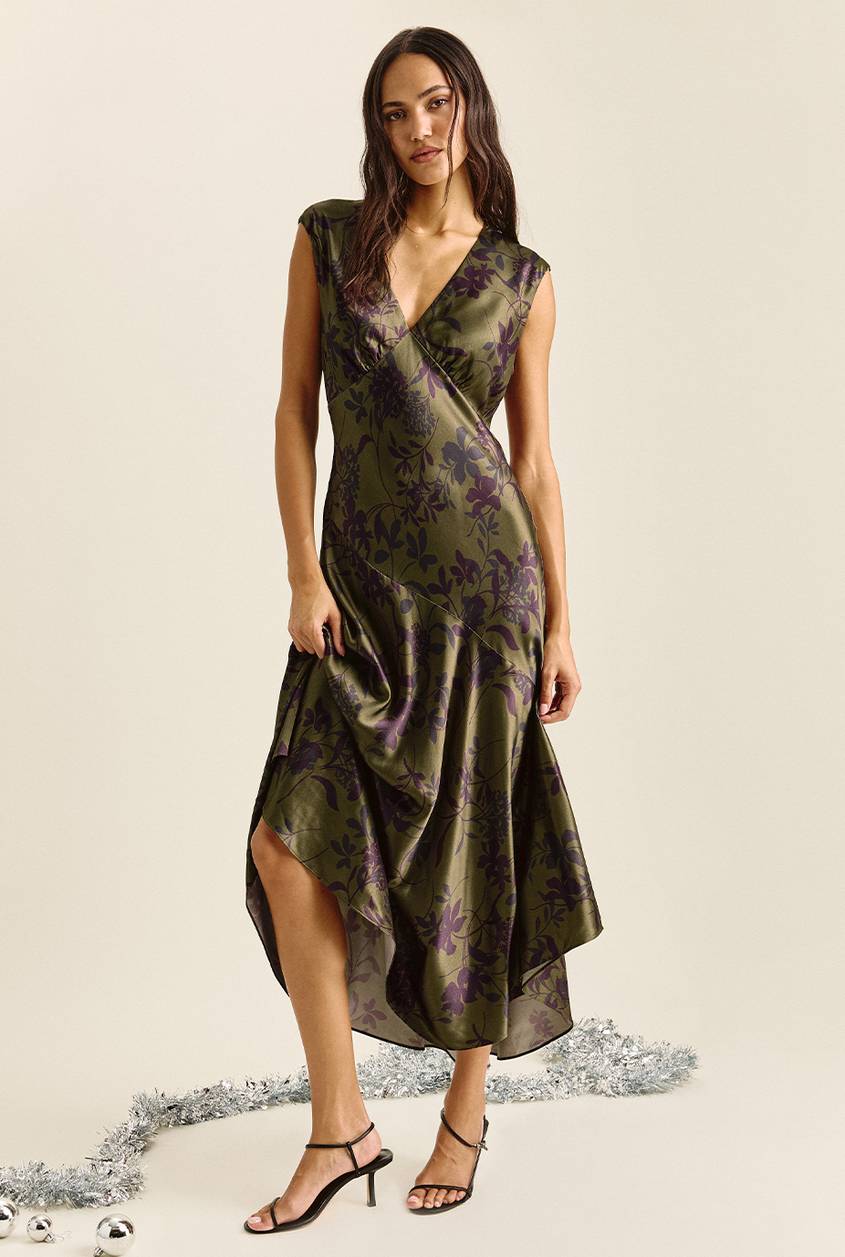 Woman standing and holding the skirt of a sleeveless olive green satin midi dress with a deep V-neck, a purple floral print, and an asymmetrical hem, paired with black strappy heels. Silver tinsel and ornaments lie on the floor.