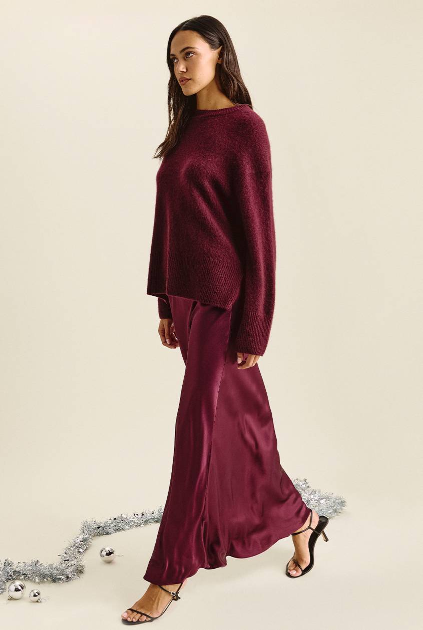 Woman walking and looking to the side, wearing an oversized burgundy sweater over a matching burgundy satin maxi skirt, with black strappy heels. Silver tinsel and ornaments lie on the floor.