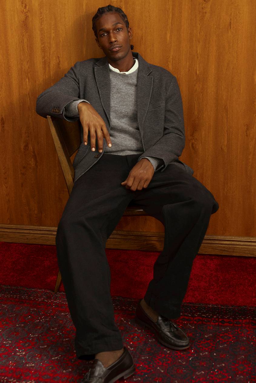 Man seated in a wooden chair, wearing a charcoal gray knit blazer over a gray crewneck sweater and white collared shirt, with black wide-leg trousers and dark brown tassel loafers. A red patterned rug is on the floor.