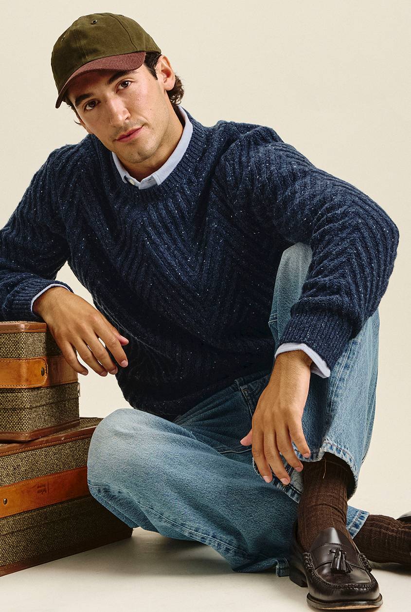 Man seated on the floor in an olive and burgundy baseball cap, a navy speckled chevron-knit sweater over a light blue collared shirt, light-wash jeans, brown ribbed socks, and dark tassel loafers. He leans on a stack of tweed suitcases.