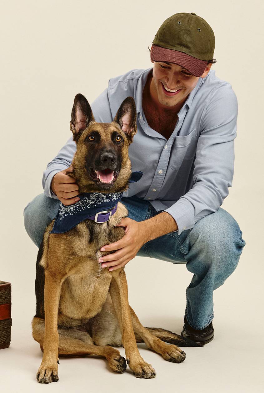 Smiling man crouching beside a German Shepherd, wearing an olive and burgundy baseball cap, a light blue button-up shirt, and light-wash jeans. The dog wears a navy paisley bandana and a purple collar.