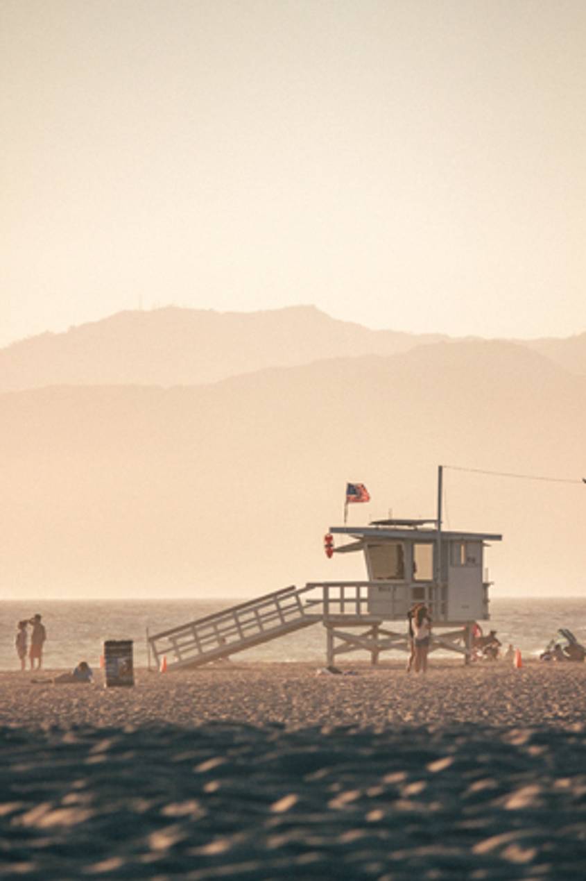 A wooden lifeguard tower with an American flag on a sandy beach, overlooking the ocean at dusk with a warm, hazy sky.