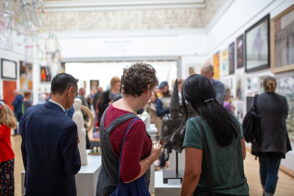 Three people facing away are browsing an art exhibition with artworks on the walls and people in the distance 