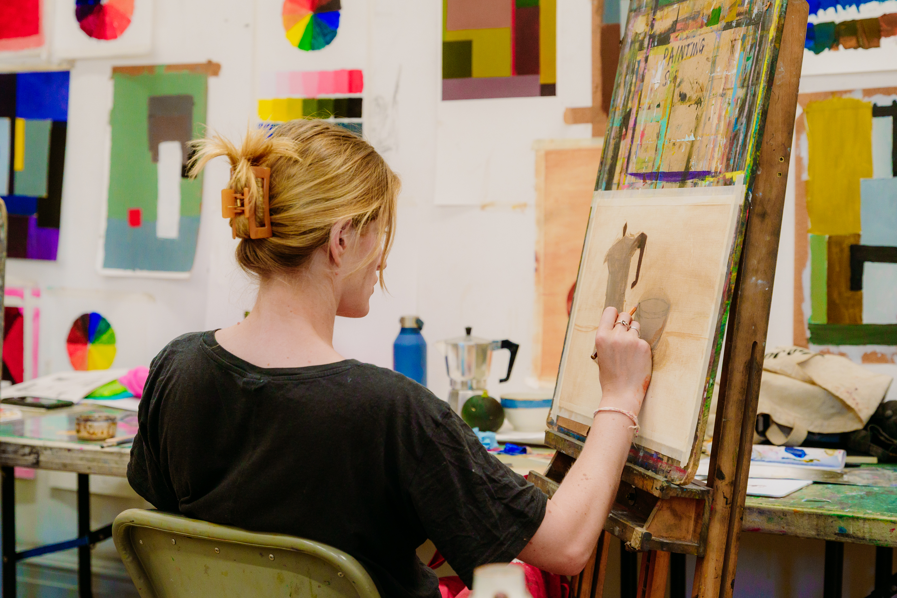 Back and side profile of a blond white woman at an easel painting with paintings on the wall in front of her 