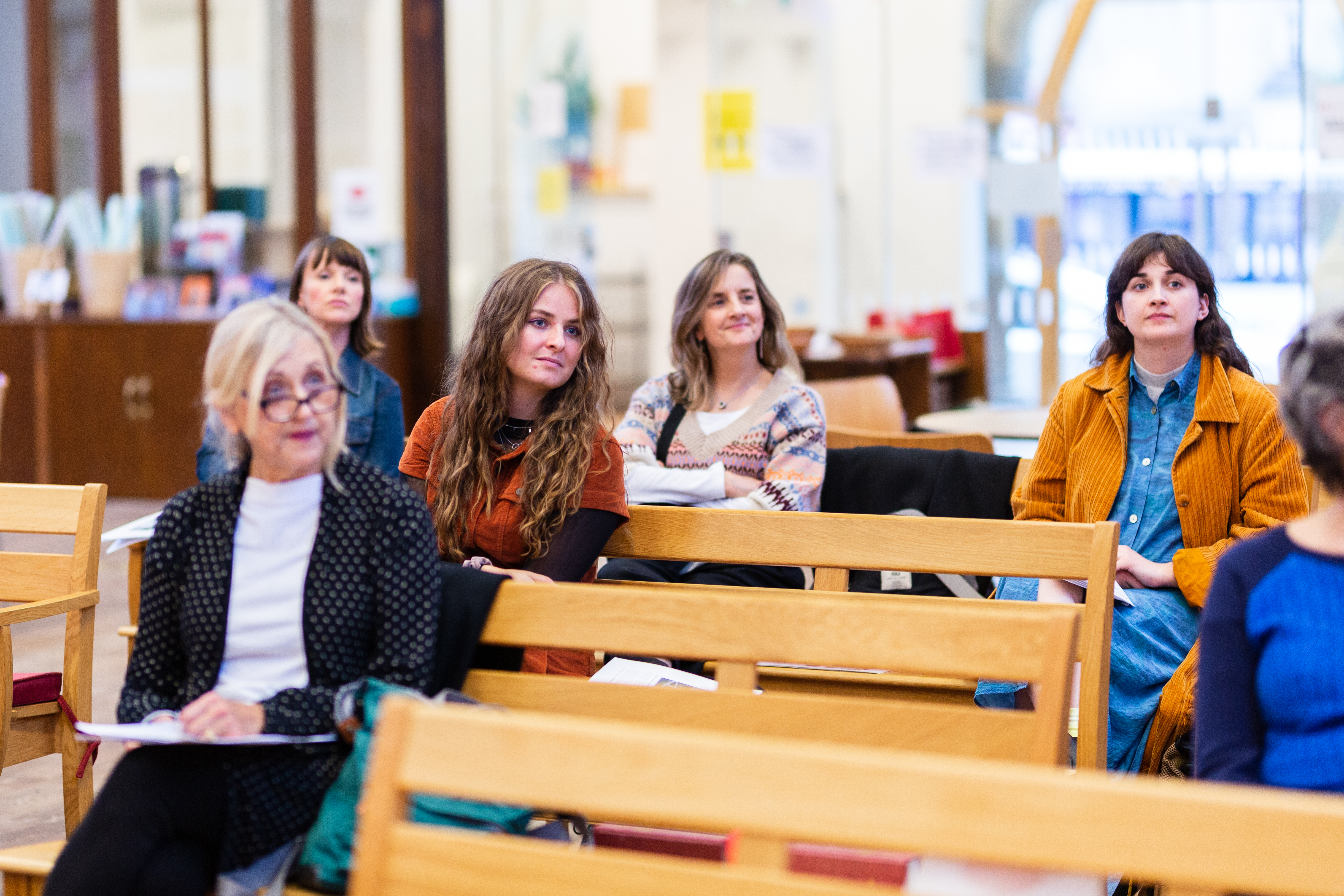 5 women sat on benches spreadout looking ahead to the right 