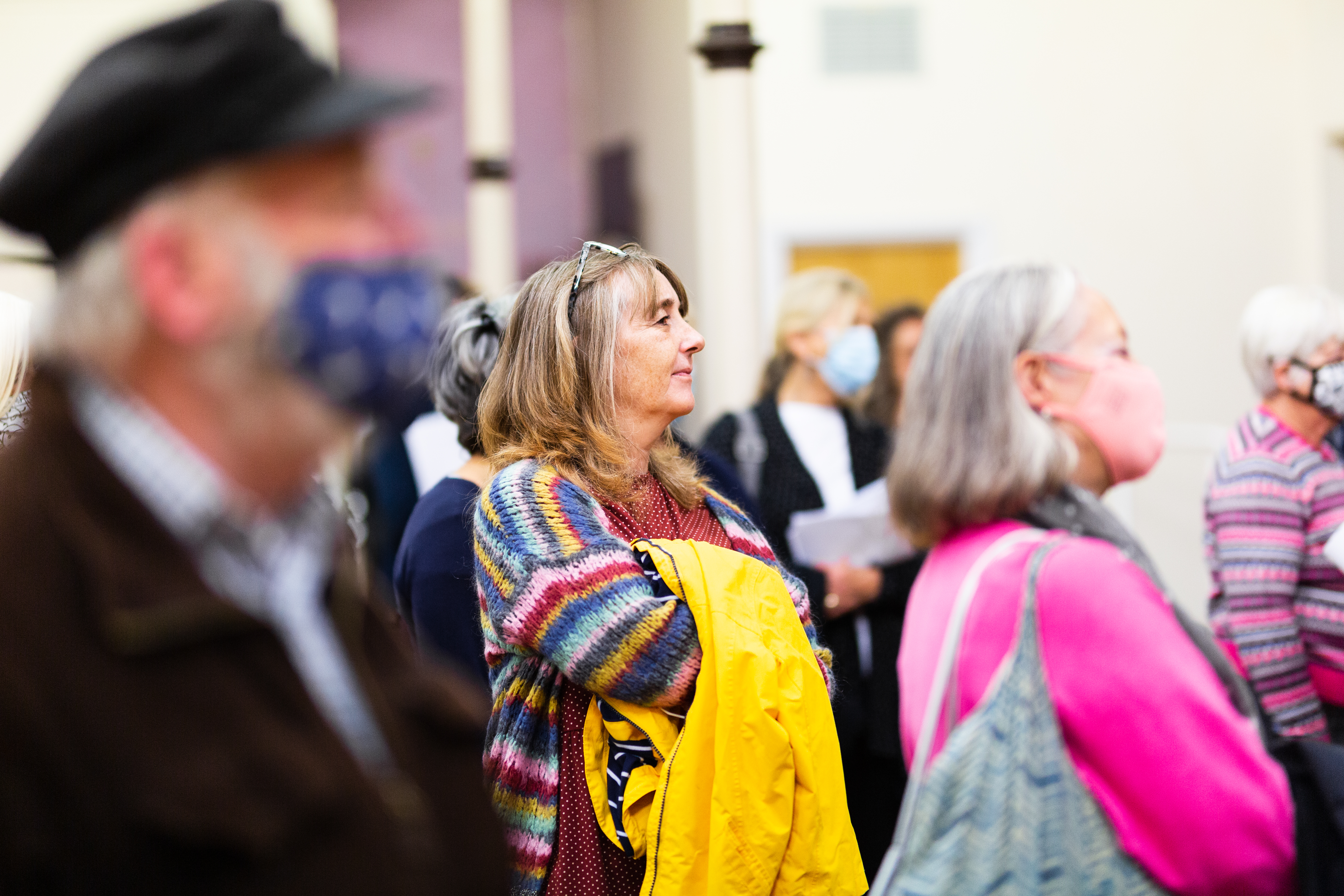 Focusing on one white, grey haired woman in the middle, with her arms crossed holding a yellow coat. Surrounded by other adults all facing to the right that are out of focus 