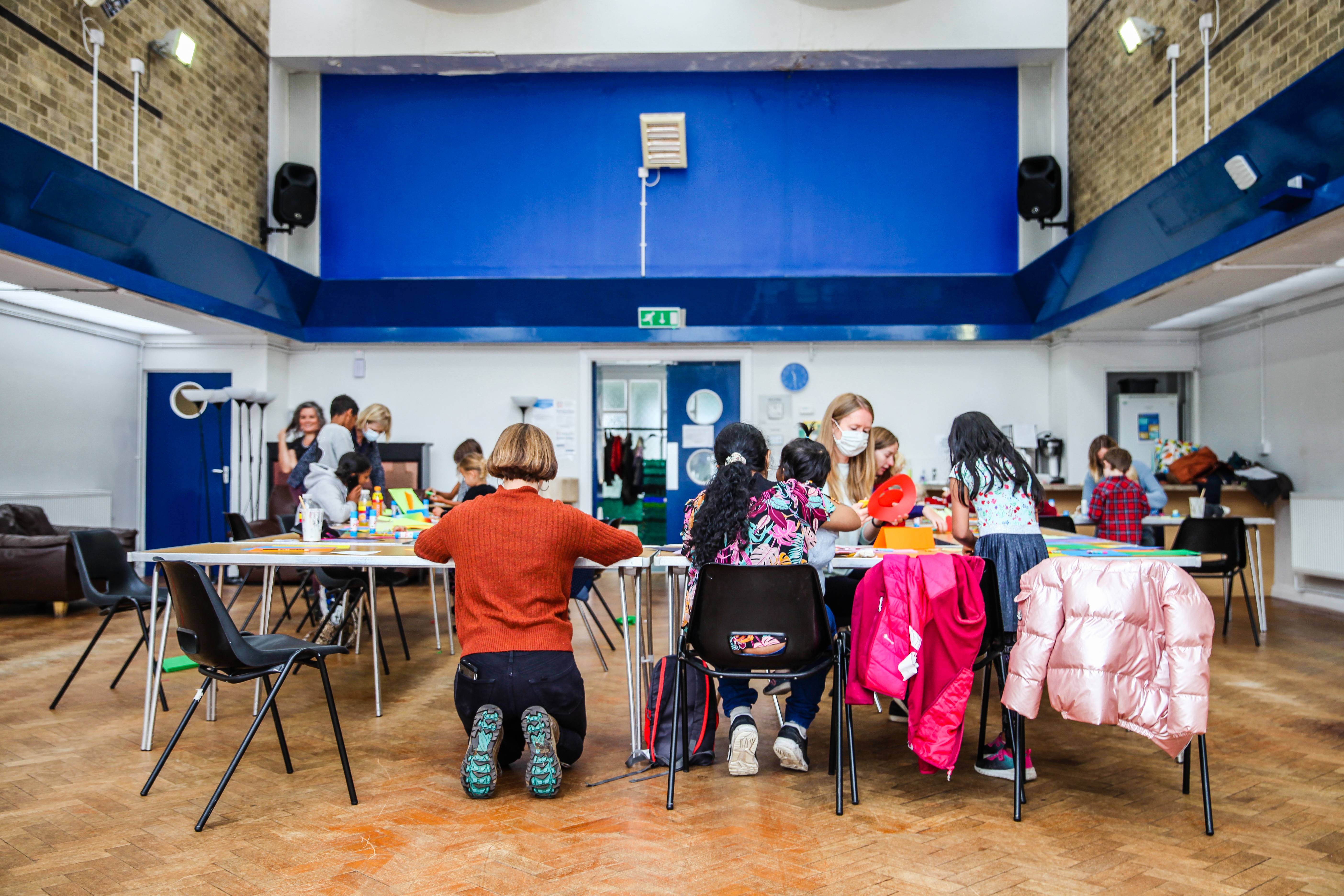 Groups of young children sat at tables. The back of an adult knelt at the table in the forground 