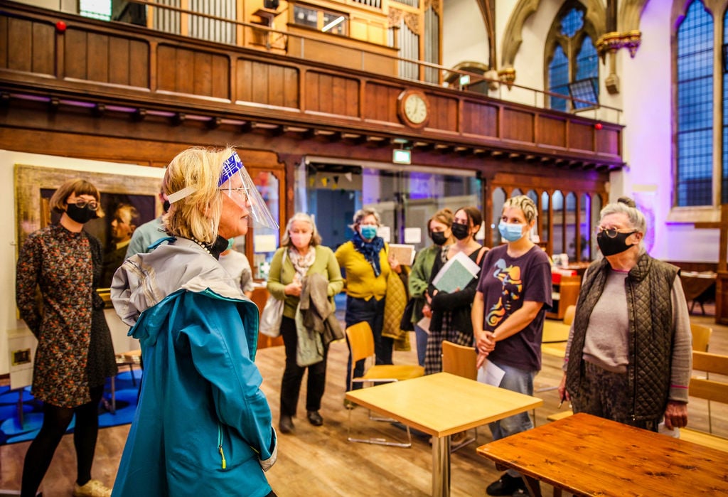 A semi circle of adults standing facing forward, wearing face masks. One woman in a visor stood in front of them to the left, facing right. They are stood with two tables in front of them on the right and the the interior of the church wooden balcony above them 