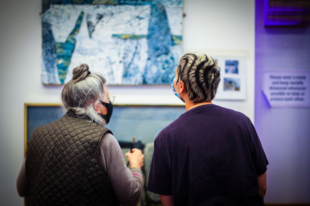 A blue abstract painting in the background, with the backs of two women facing the painting in the foreground 