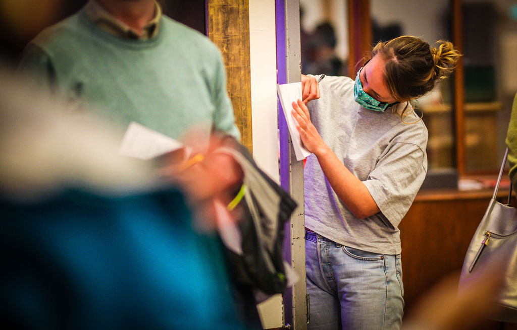 In focus on the right is a young adult, facing forward to the left, using the wall to the left to write notes. Out of focus on the left is an adults torso and hands 